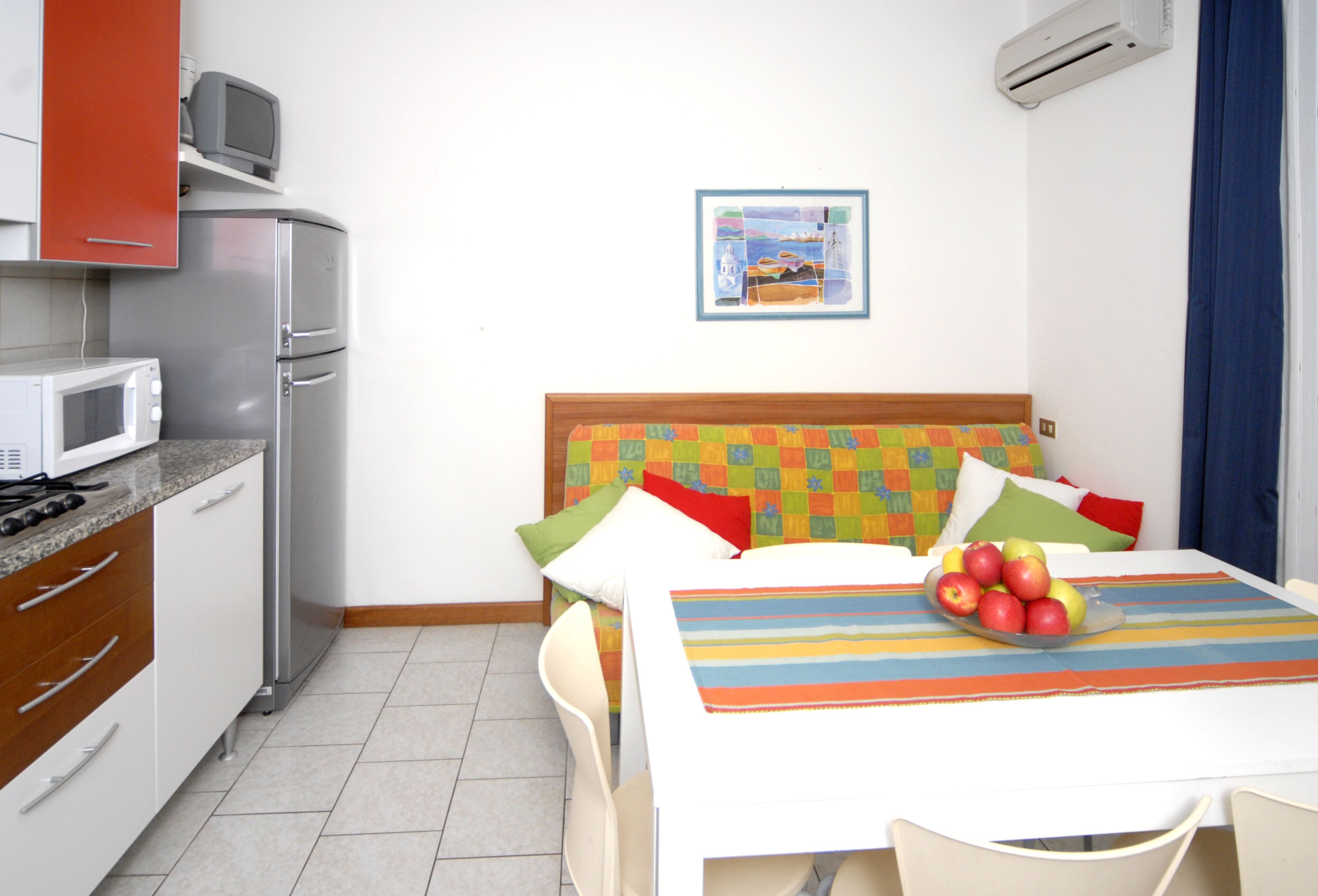 Camera d'albergo compatta con angolo cottura, un letto con biancheria colorata, un quadro appeso al muro e un tavolo con una ciotola di frutta.