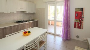 A modern kitchen with white cabinets, a gas stove, and a dining area with a white table and chairs. - Modern one Bedroom Flat in Bibione - Beahost (San Michele al Tagliamento)