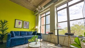 Una stanza con un divano blu, un tavolino da caffè in vetro, un tappeto verde e ampie finestre che offrono vista su edifici e alberi.