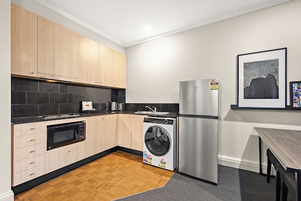 A compact kitchen with wooden cabinets, a stainless steel refrigerator, a built-in washer and dryer, a microwave, and a small dining table. - Melbourne City Suites (Melbourne)