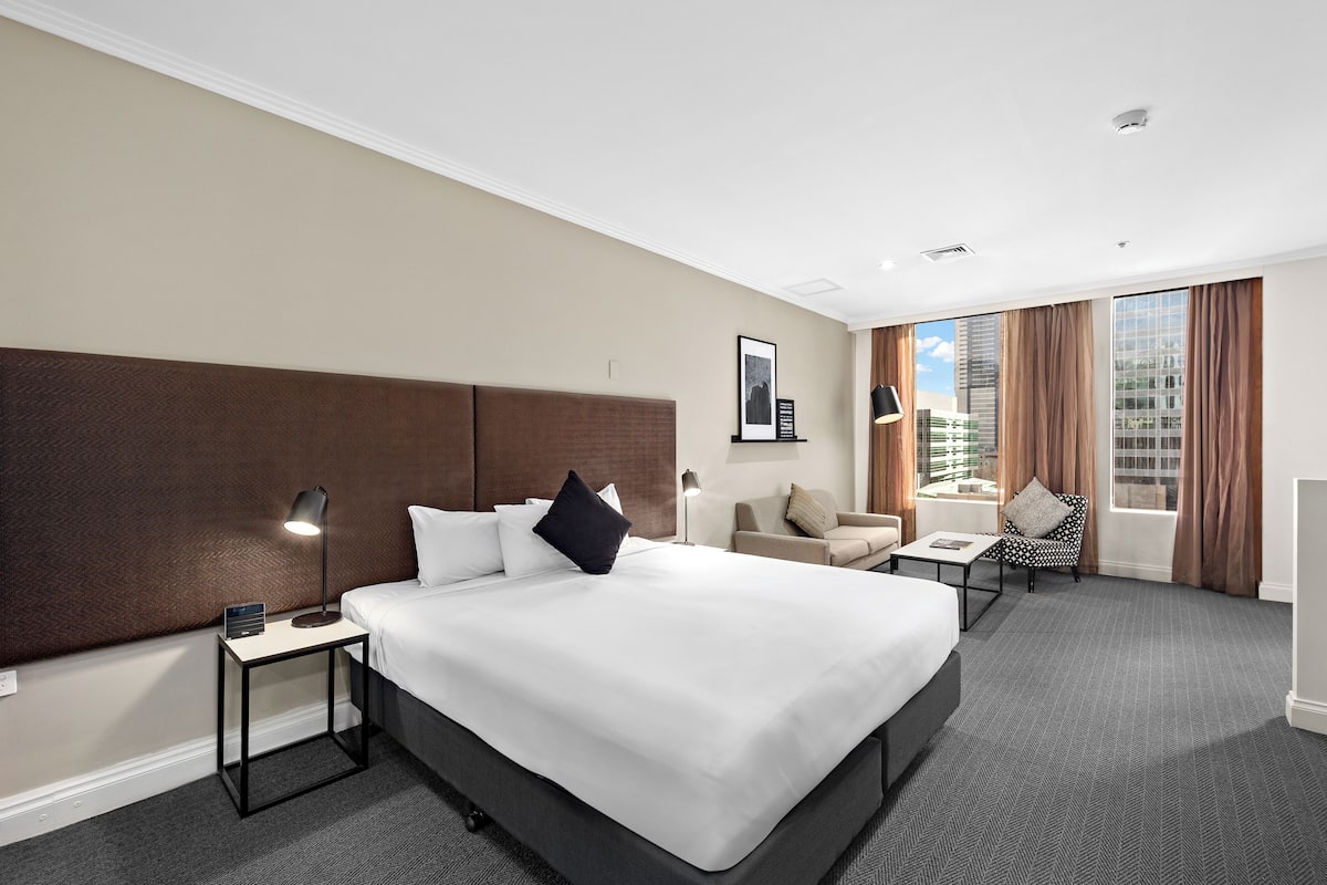 Ein Hotelzimmer mit einem großen Bett, einem Nachttisch mit Lampe, einer kleinen Couch und einem Fenster mit Vorhängen.