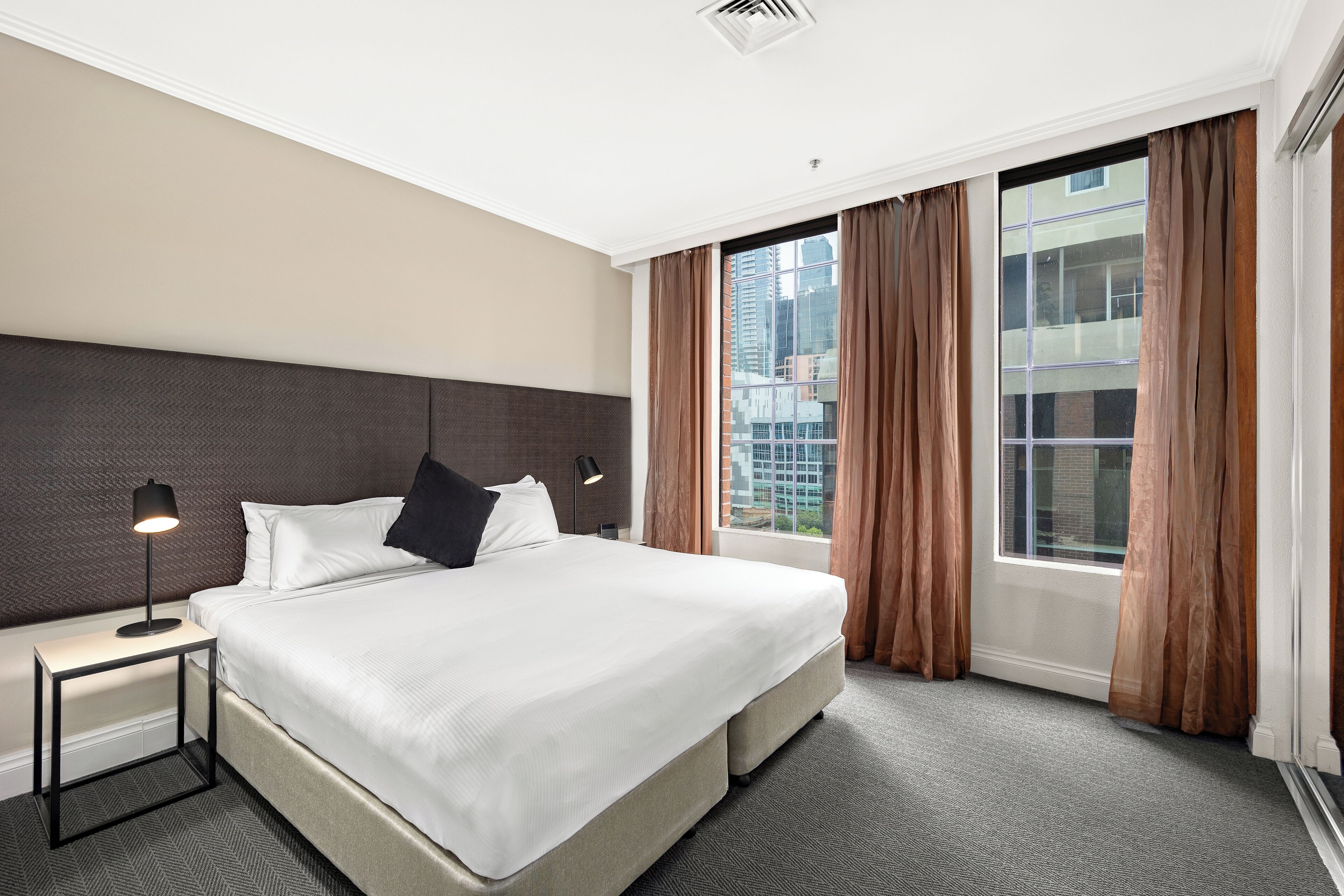 A hotel room with a large bed, a nightstand with a lamp, and a view of a cityscape through large windows.