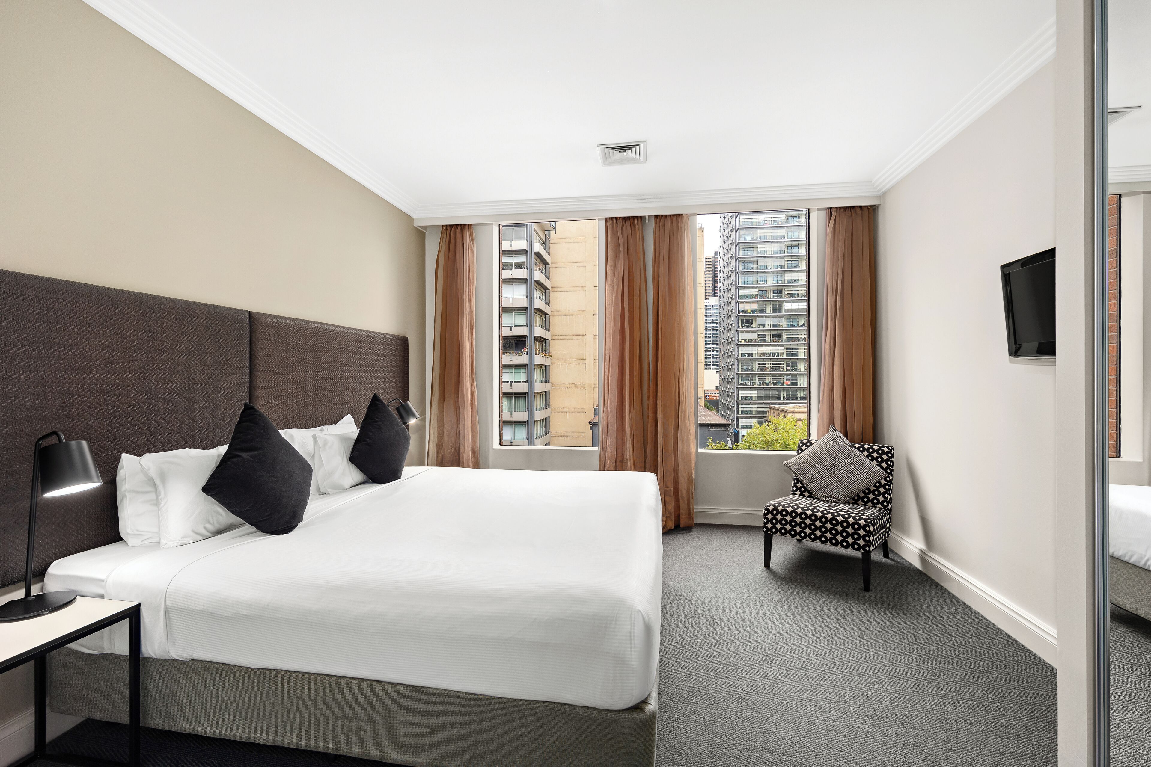 A hotel room with a large bed, a desk with a lamp, a chair, and a TV on the wall.