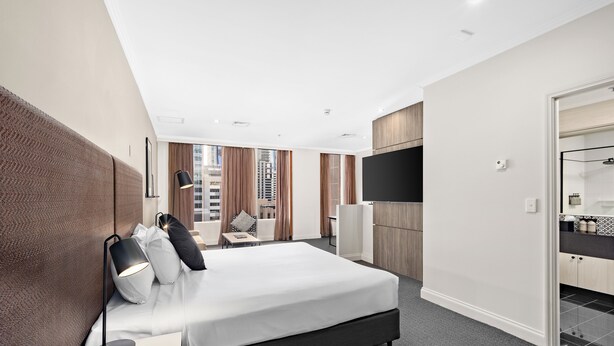 Una habitación de hotel moderna con una cama grande, un escritorio con lámpara, televisión y un baño visible a través de una puerta abierta.