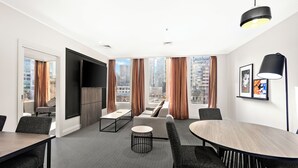 Una habitación de hotel moderna con un amplio ventanal que ofrece vista a la ciudad, un televisor de pantalla plana, una mesa de comedor redonda y una zona de estar confortable.