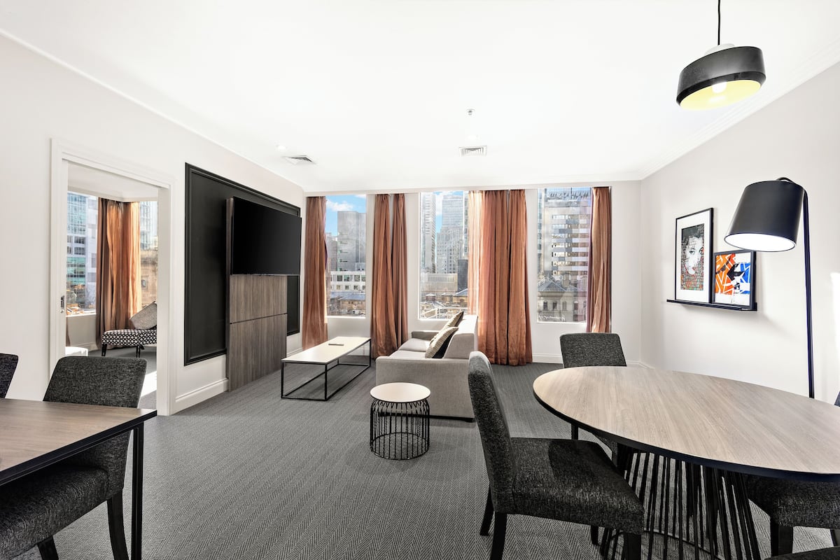 Ein modernes Hotelzimmer mit großem Fenster und Blick auf die Stadt, einem Flachbildfernseher, einem runden Esstisch und einer gemütlichen Sitzecke.