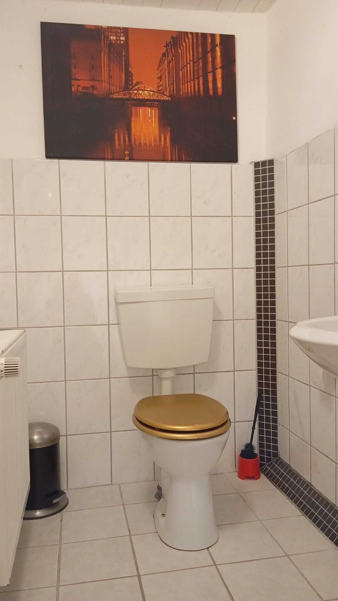 A small bathroom with a white toilet, a wooden seat, a tiled wall, a black and white tile accent, a white sink, and a trash can.