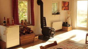 Une pièce chaleureuse avec une cheminée, un petit réfrigérateur, un panier en osier rempli de bois de chauffage et une fenêtre avec des rideaux.
