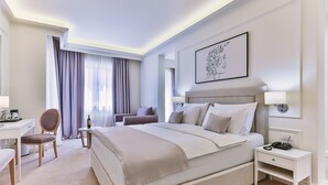 A hotel room with a large bed, a desk, a chair, a small table, and a nightstand with a phone. - Hotel Crown (Budva)
