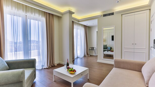 A modern living room with a sofa, a coffee table with fruit, and a view of a cityscape through large windows. - Hotel Crown (Budva)