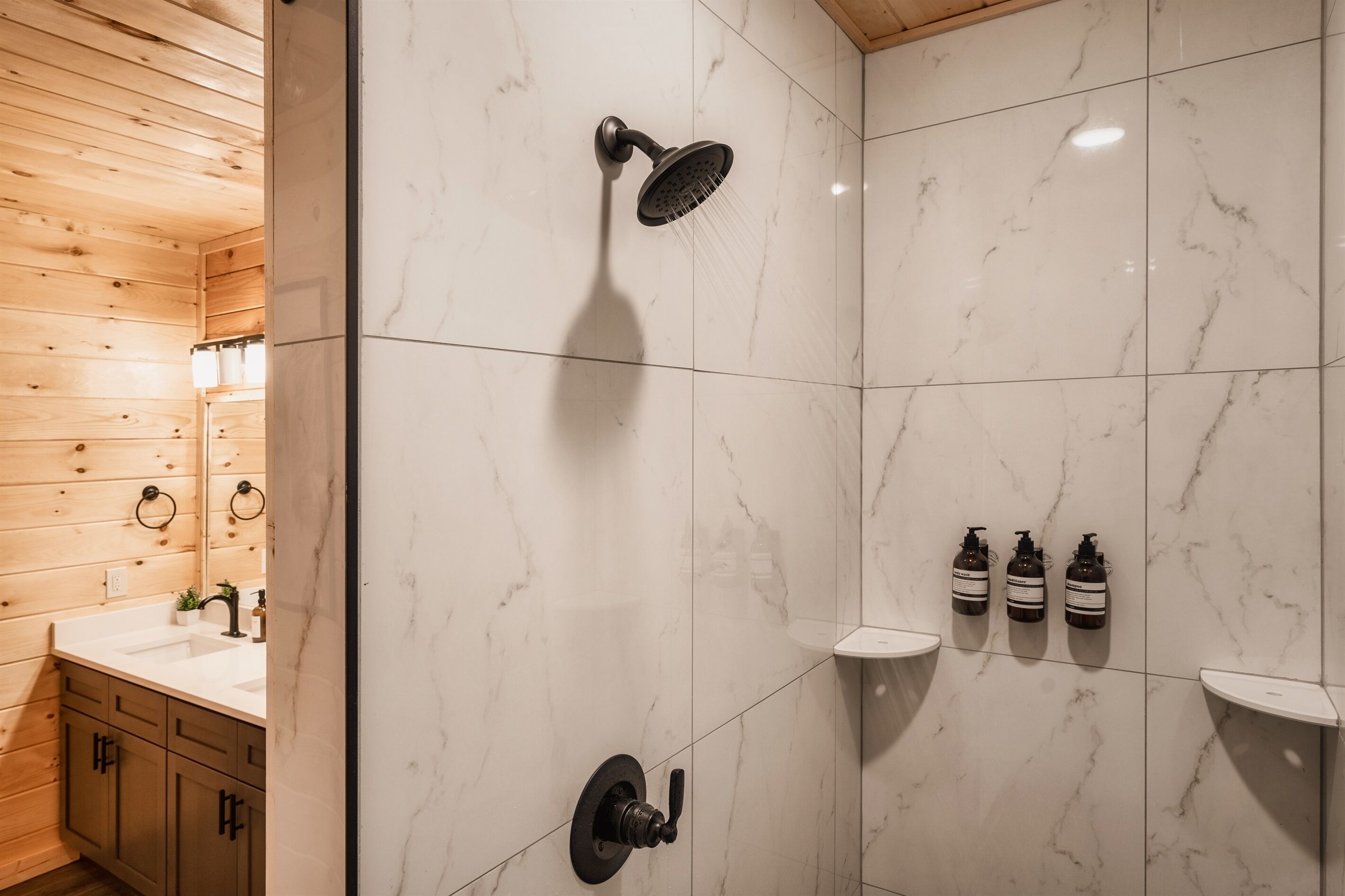 Moderni kylpyhuone, jossa on suihku, pesuallas ja hyllyllä kolme pulloa shampoota.