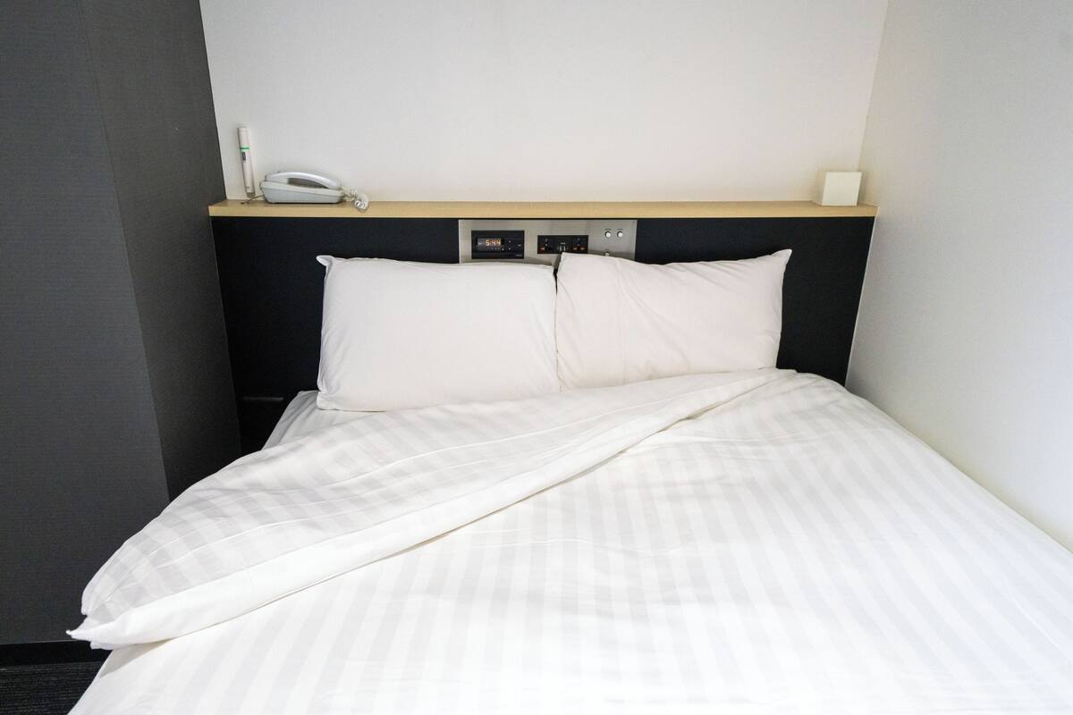 Ein ordentlich bezogenes Bett mit weißen Leinen, ein Nachttisch mit Lampe und eine an der Wand montierte Steuerung.