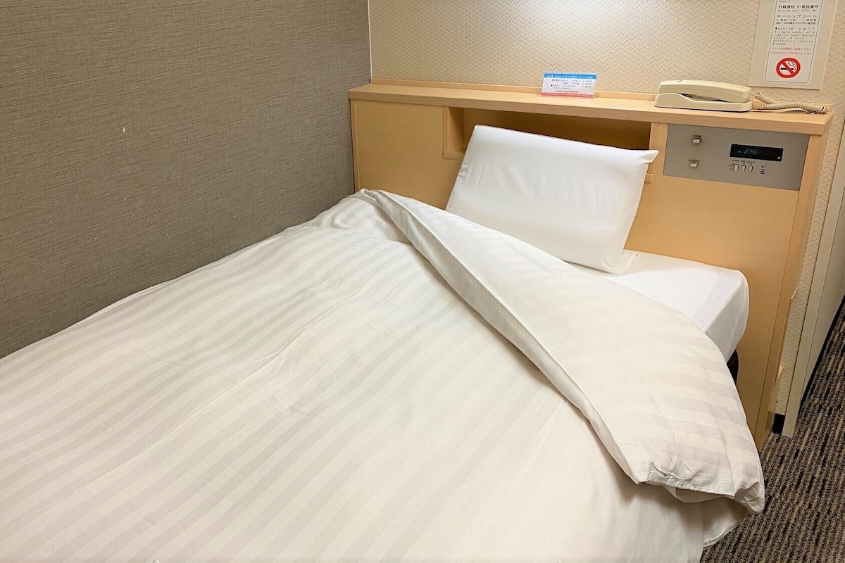 Un lit simple avec une literie blanche, une tête de lit en bois, une table de chevet avec un téléphone et une horloge numérique.