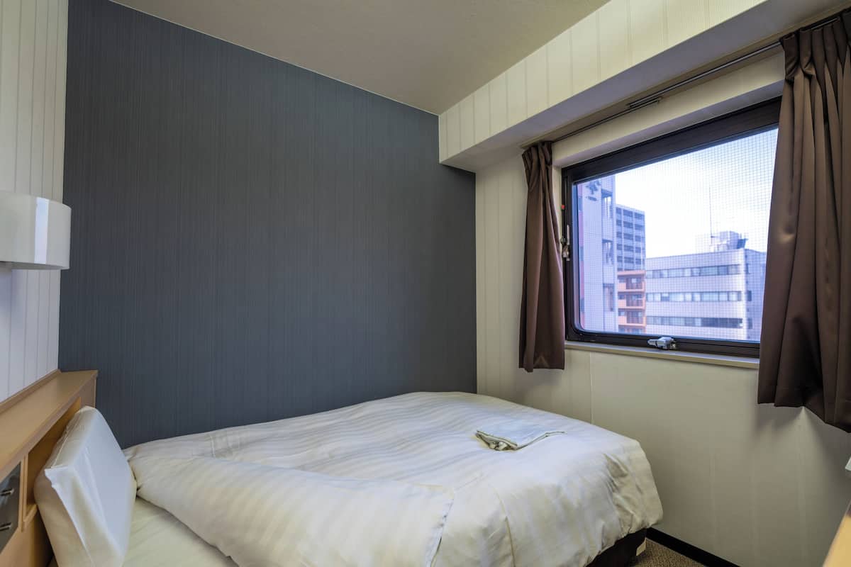 Ein Hotelzimmer mit Bett, einem fensterbelaubten Fenster, einer Wandleuchte und Blick auf umliegende Gebäude.