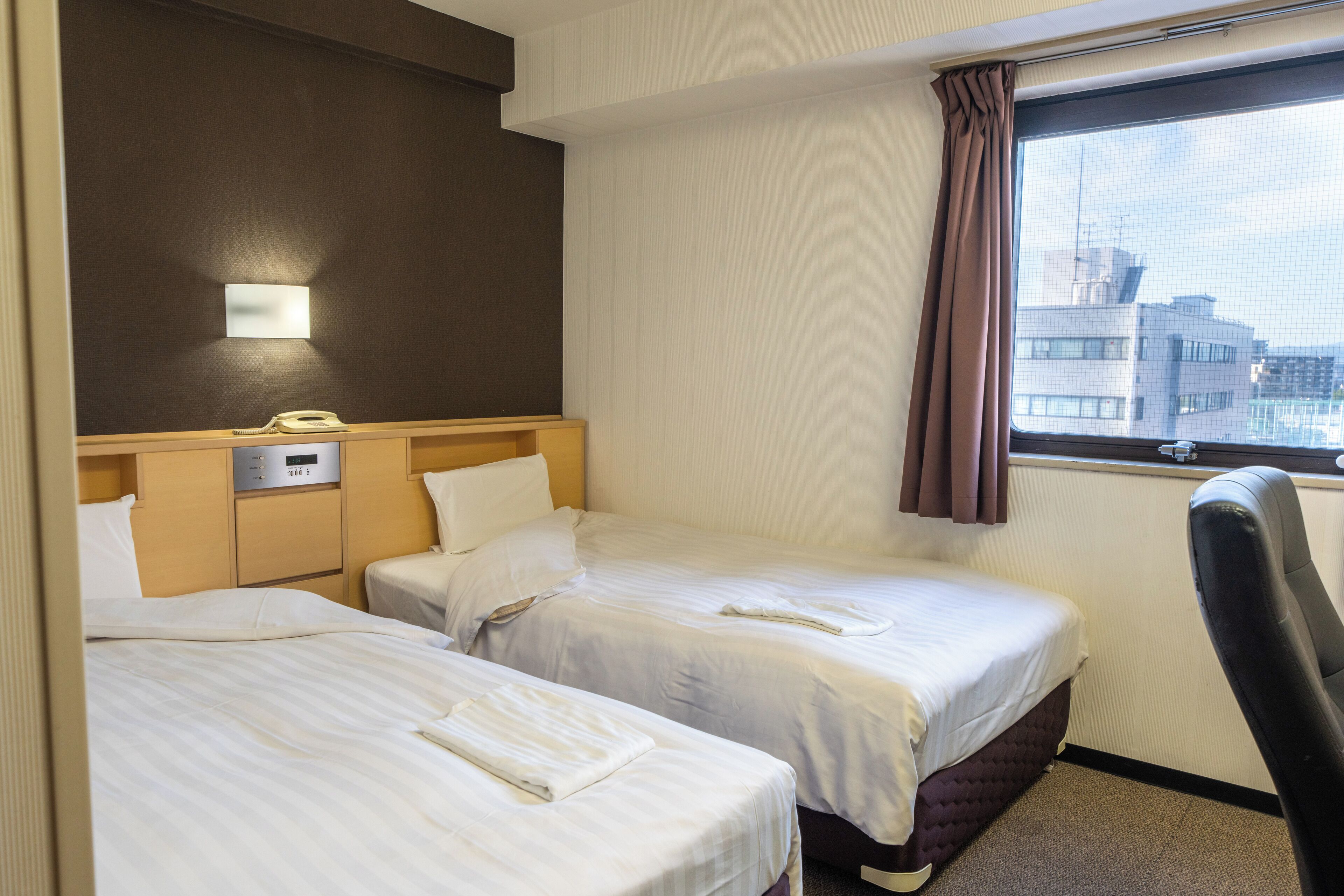 A hotel room with two beds, a desk with a telephone, a window with curtains, and a chair.