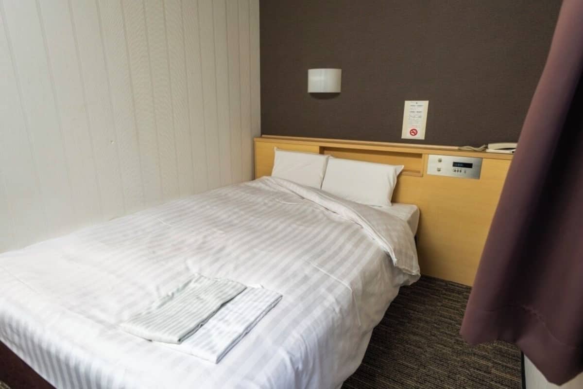 Ein Hotelzimmer mit einem ordentlich bezogenen Bett, einer Wandlampe und einer Bedienkonsole an der Wand.