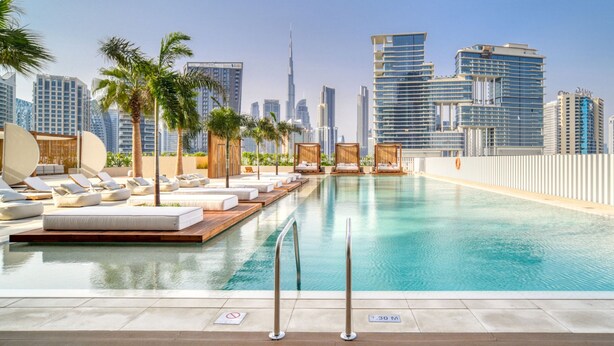 A rooftop infinity pool with lounge chairs and palm trees, overlooking a city skyline with modern buildings. - Modern Studio w Balcony in Business Bay (Dubai)