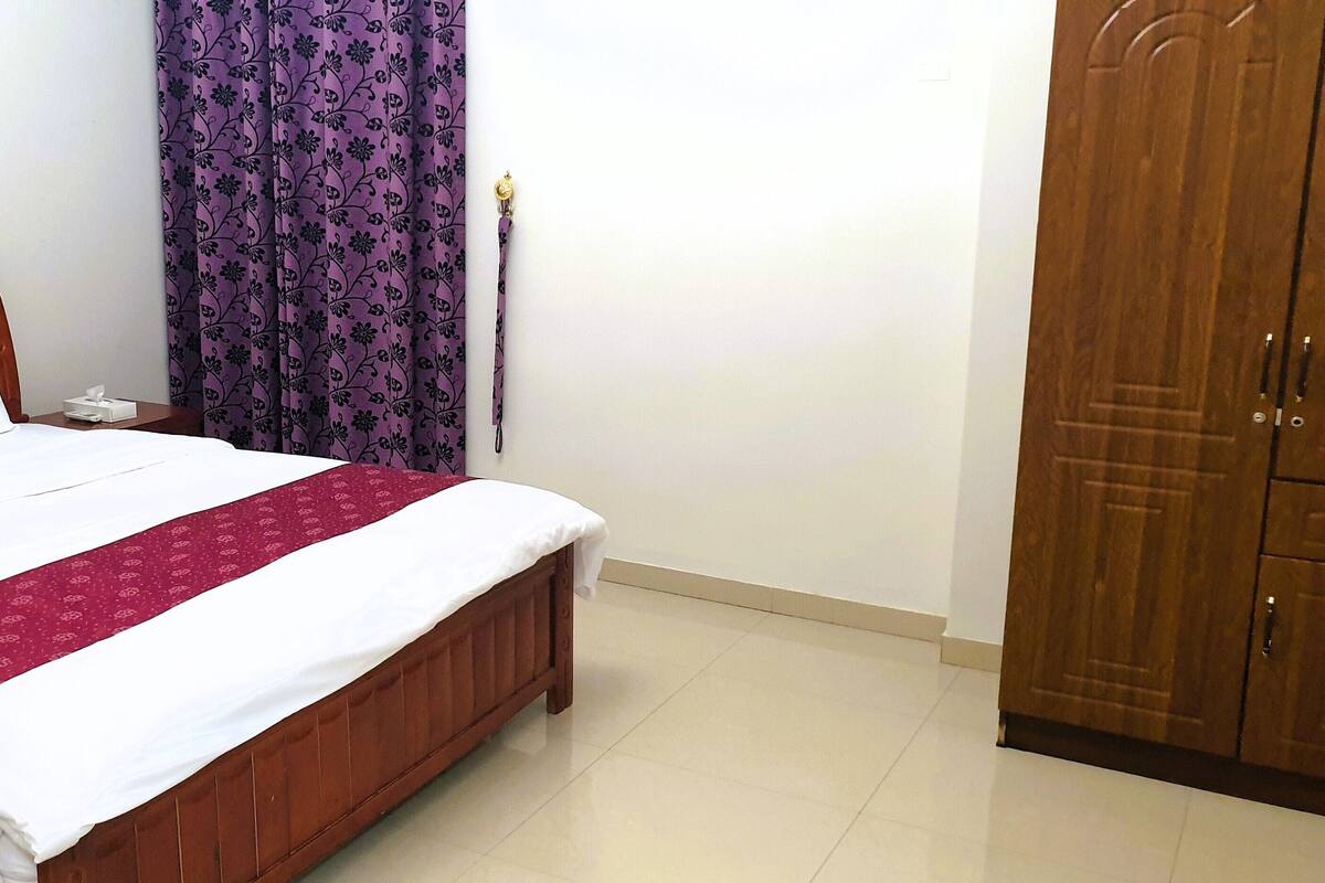 A neatly made bed with white and red linens, a wooden headboard, a nightstand with a lamp, and a purple curtain.