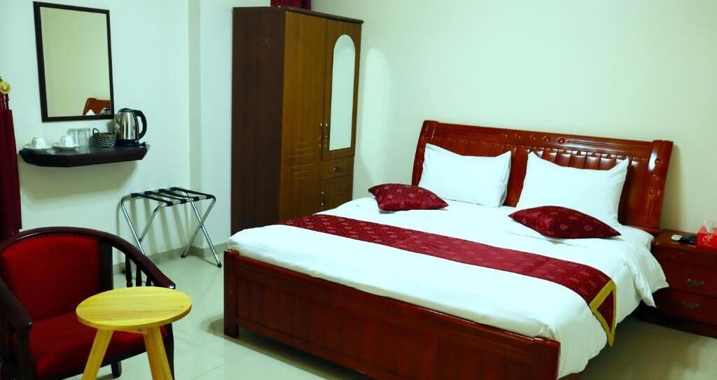 Una habitació d'hotel amb un llit de fusta, dos coixins, un llençol vermell, un armari de fusta, una taula petita, una cadira i un mirall a la paret.