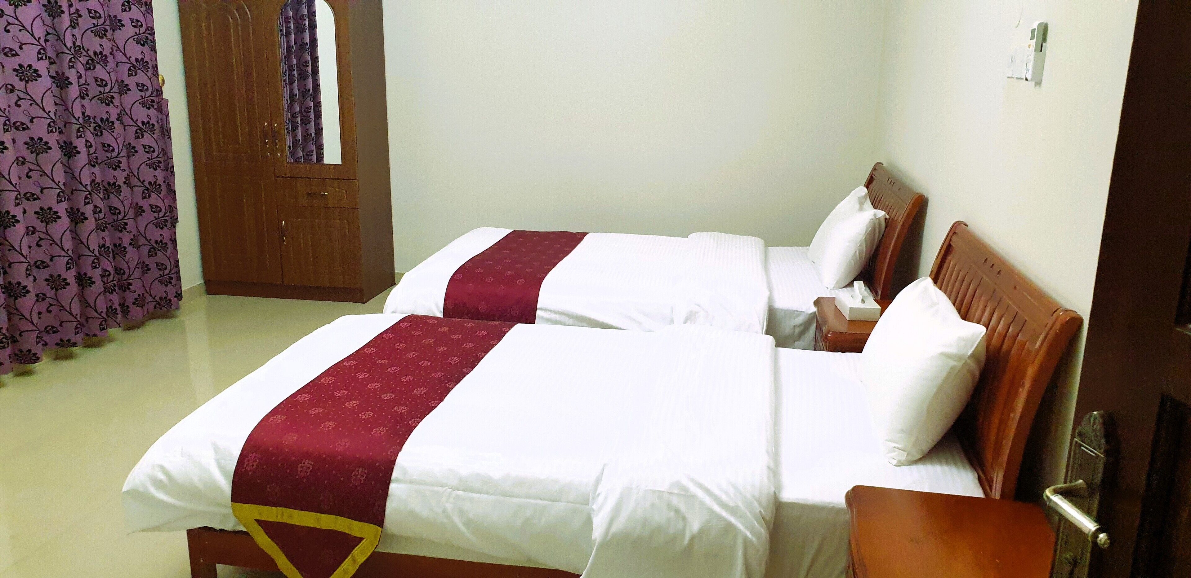 A hotel room with two beds, each with white linens and a red decorative strip. There is a wooden door with a mirror and a purple curtain with a floral pattern.