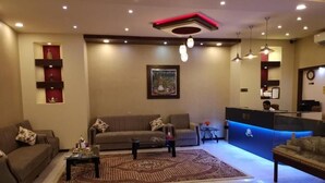 A modern living room with a sofa set, a coffee table, and a patterned rug. There is a reception desk with a blue accent wall and a clock on the wall. - Bahla Hotel Apartment  (Bahla)