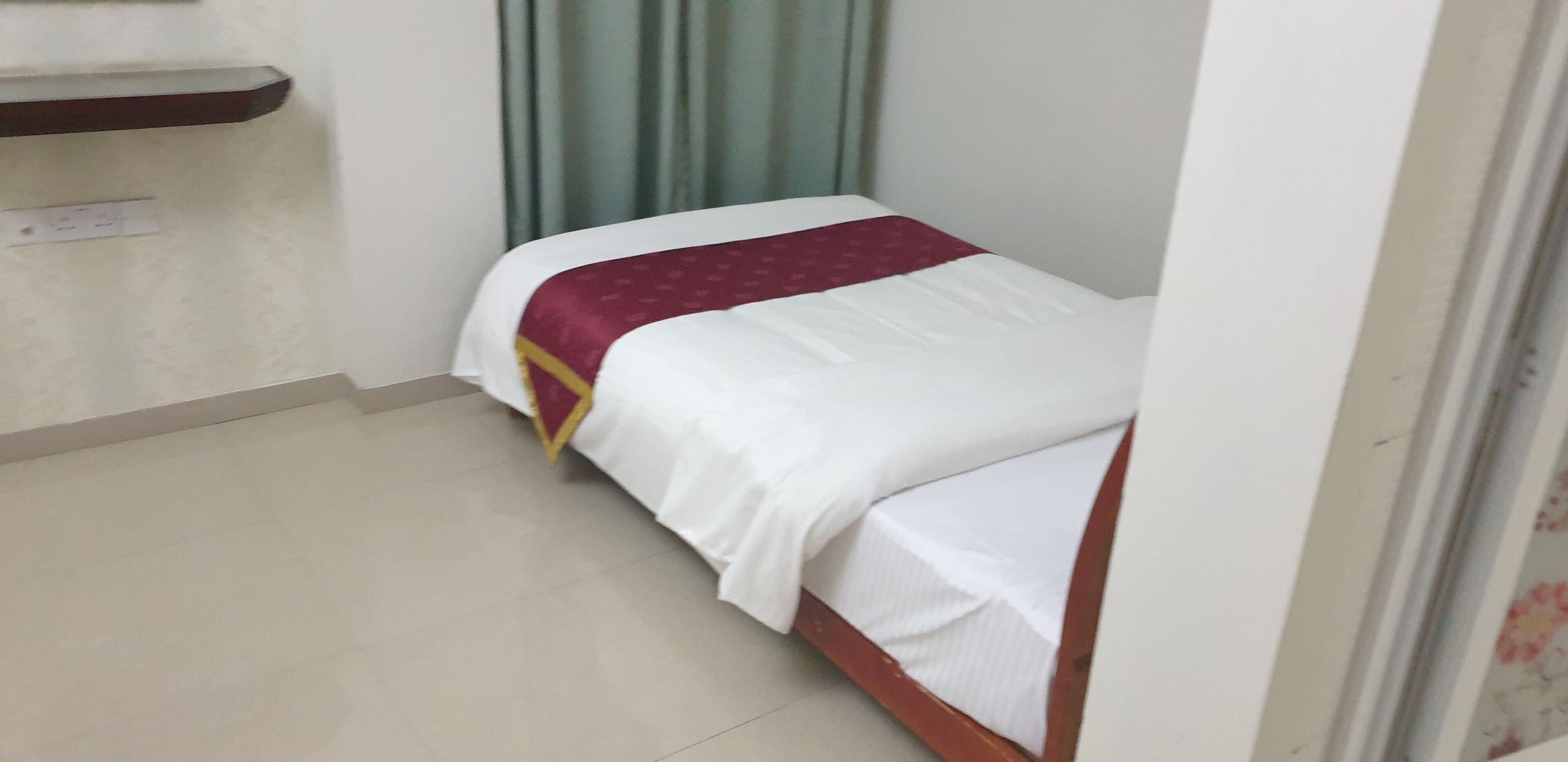 A small, single-bed room with a wooden frame, white bedding, and a maroon accent pillow.