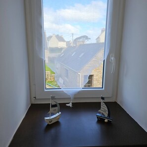 Una ventana con dos pequeños veleros en un alféizar oscuro, con vistas a una zona residencial con edificios de piedra y un cielo despejado.