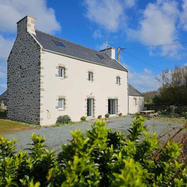 A two-story stone house with a sloped roof, multiple windows, and a small garden area with potted plants.