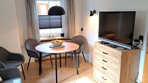 Un salon moderne comprenant une table à manger, deux chaises, un canapé, un téléviseur posé sur un meuble en bois et une fenêtre avec des rideaux.