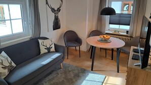 Un salon moderne avec un canapé gris, une table de salle à manger ronde entourée de chaises, un téléviseur et une grande fenêtre agrémentée de rideaux.