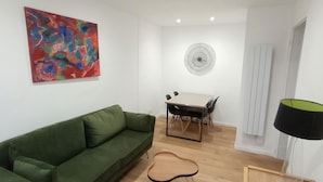 Un salon avec un canapé vert, une table à manger avec des chaises, une guitare, un tableau abstrait aux couleurs vives et une lampe.
