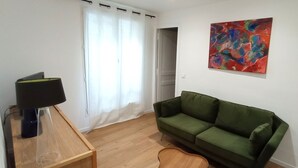 Une pièce avec un canapé vert, une petite table en bois sur laquelle se trouve une lampe, un tableau accroché au mur et une porte qui donne sur une autre pièce.
