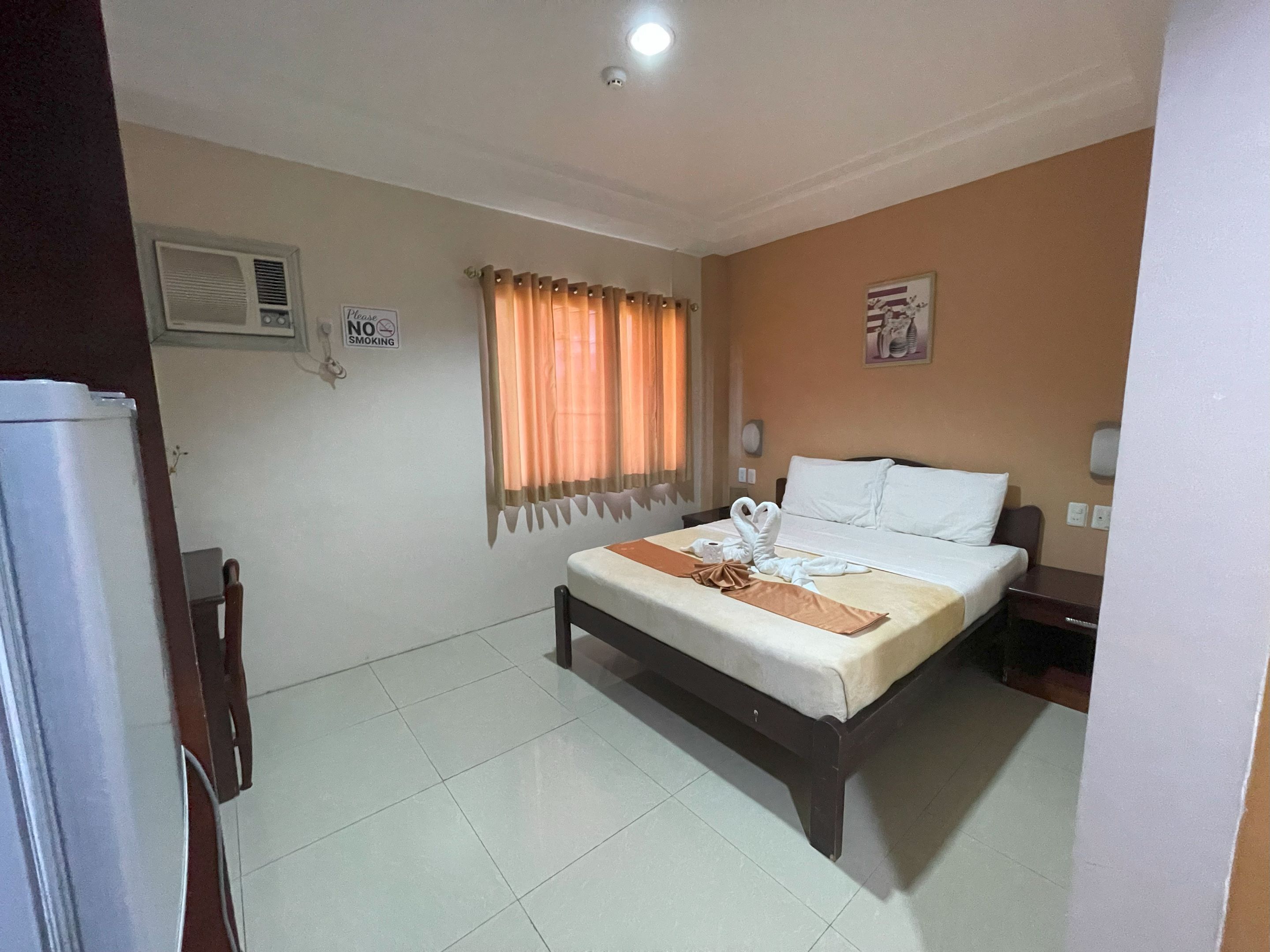 A hotel room with a bed, a bedside table, a nightstand, a window with curtains, a wall-mounted air conditioner, and a 'No Smoking' sign.
