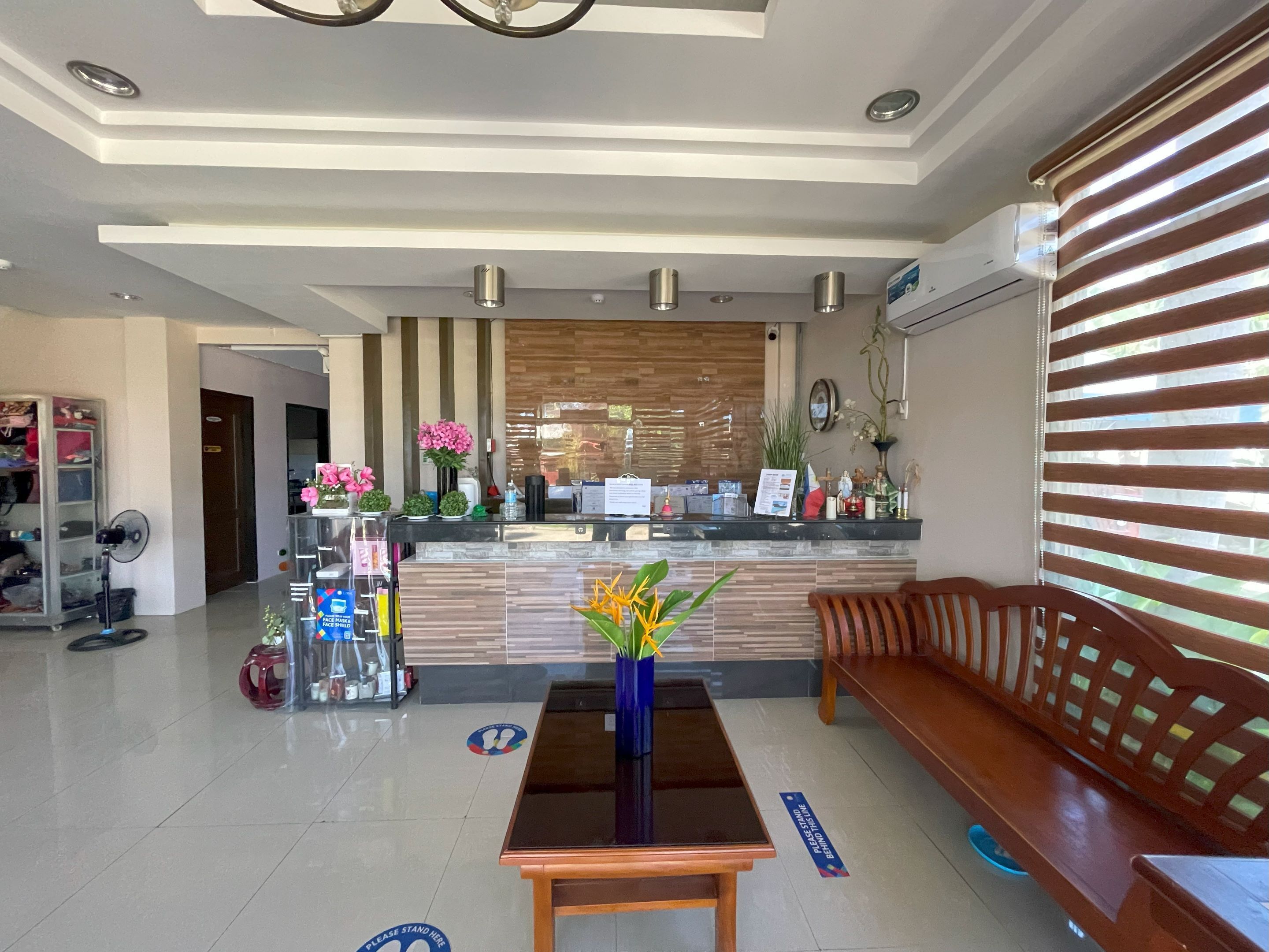 Zona de recepción de un hotel con un mostrador de madera, un banco de madera, un jarrón azul con flores y un ventilador.
