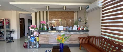 Zona de recepción de un hotel con un mostrador de madera, un banco de madera, un jarrón azul con flores y un ventilador.