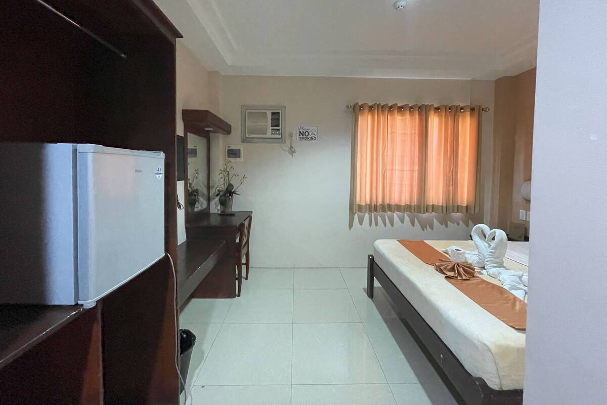 A hotel room with a bed, a desk, a refrigerator, a mirror, and a window with curtains.
