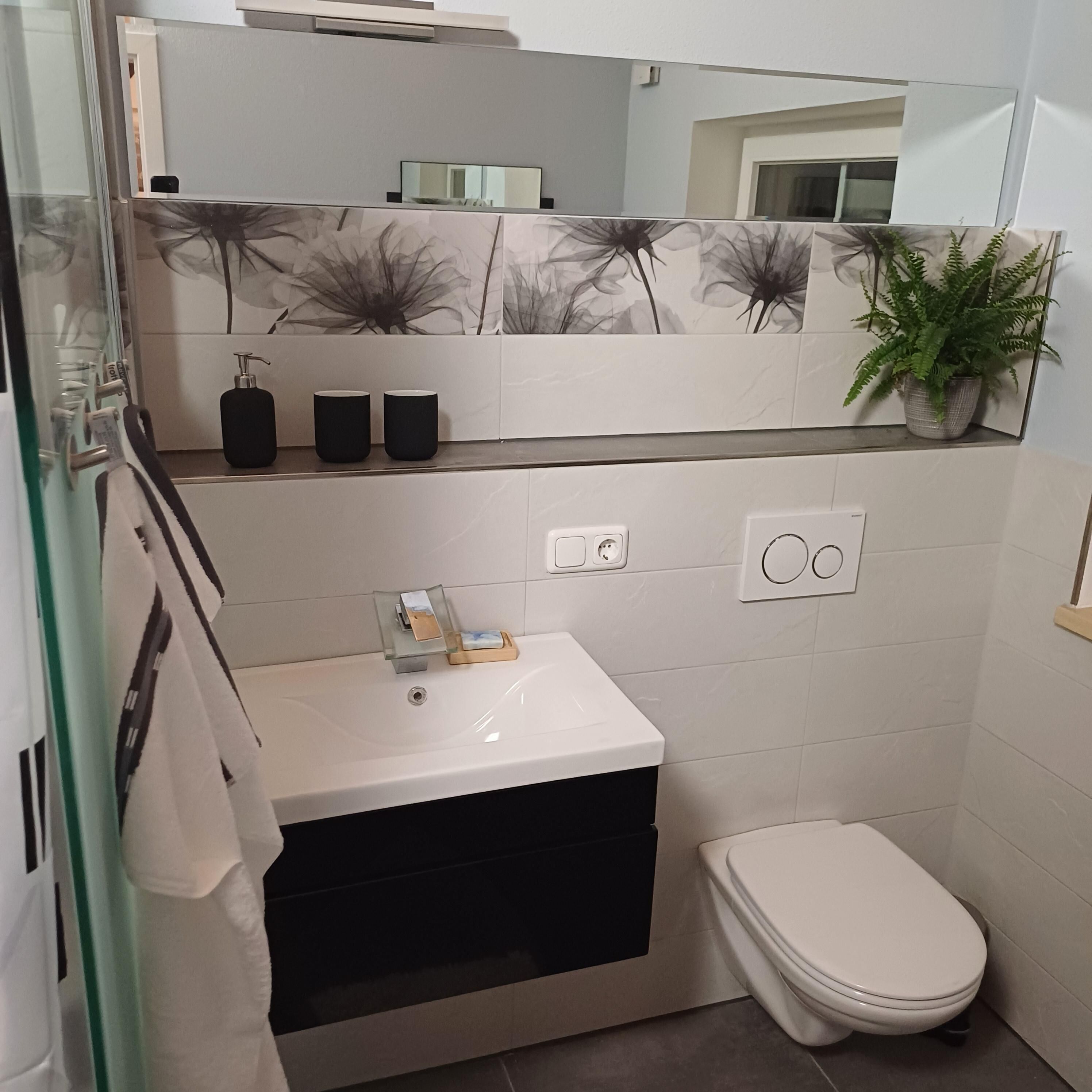 Een badkamer met een wit wastafelmeubel, een zwart kastje en een toilet. Boven het wastafelmeubel hangt een spiegel en rechts staat een plant in een pot.
