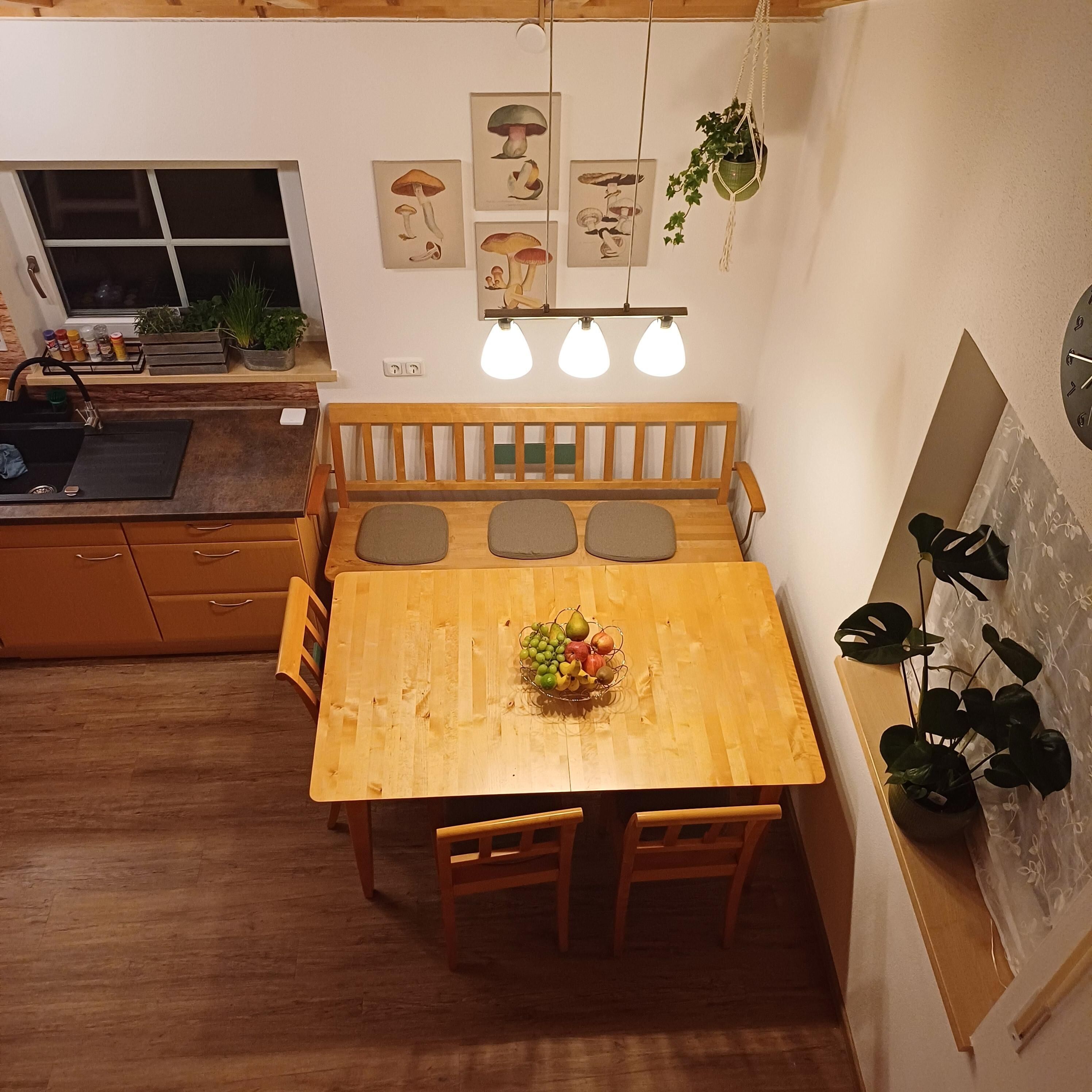 Una mesa de comedor de madera con un frutero en el centro, un banco con cojines y una lámpara colgante.
