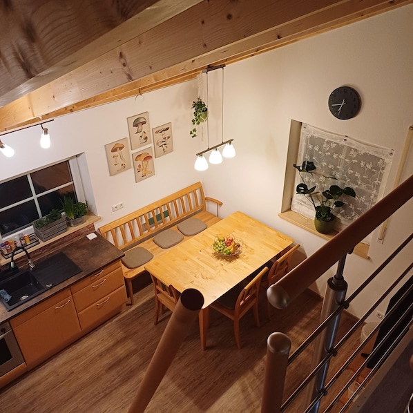 Una mesa de comedor de madera con un frutero, un banco y una zona de cocina con un lavaplatos y armarios.