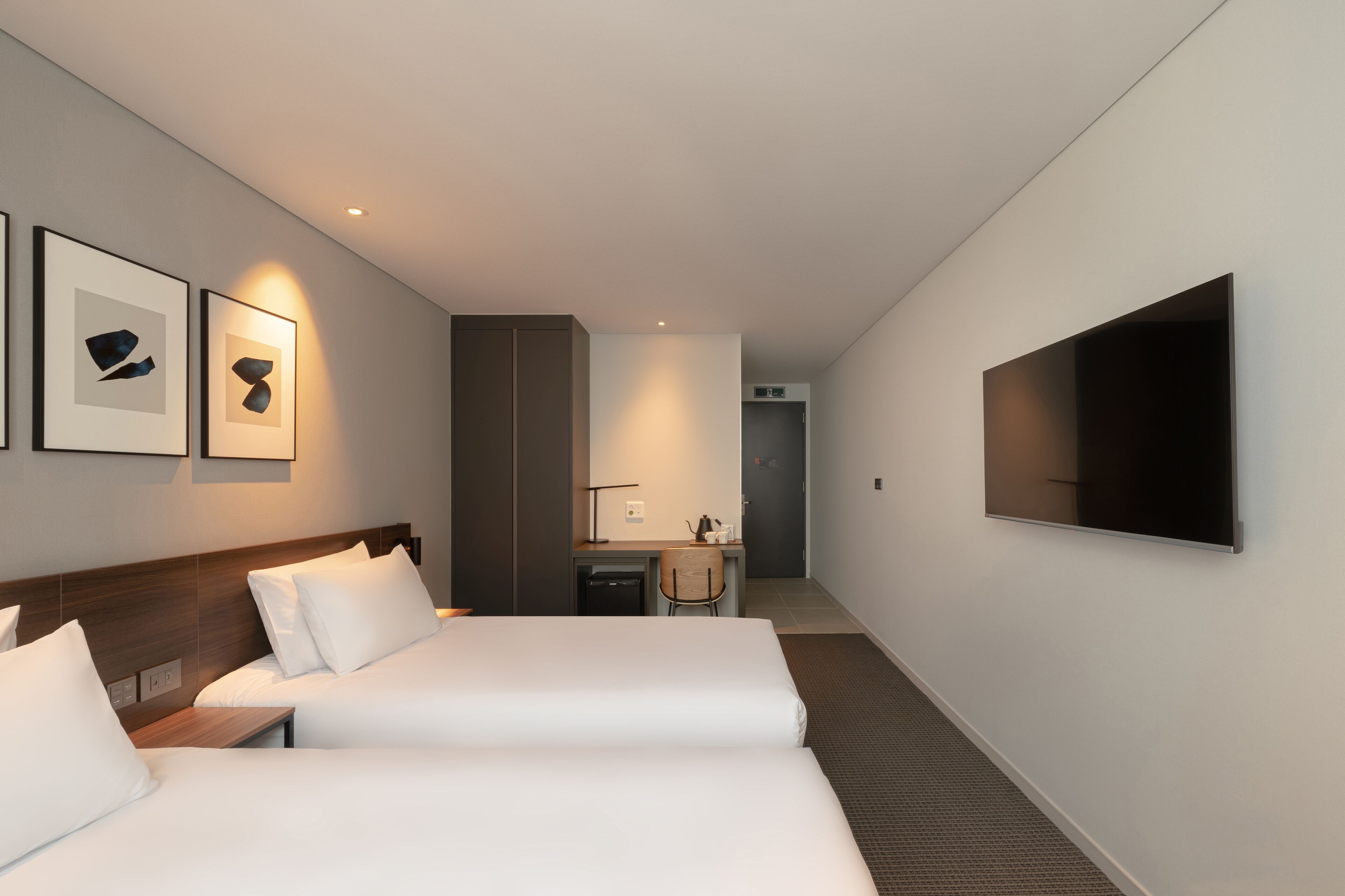 Quarto de hotel com duas camas, uma escrivaninha, uma cadeira, televisão e obras de arte na parede.