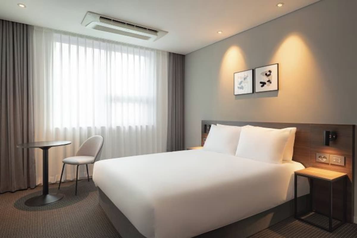 Quarto de hotel moderno com uma cama grande, uma mesa redonda, uma cadeira e uma obra de arte na parede.
