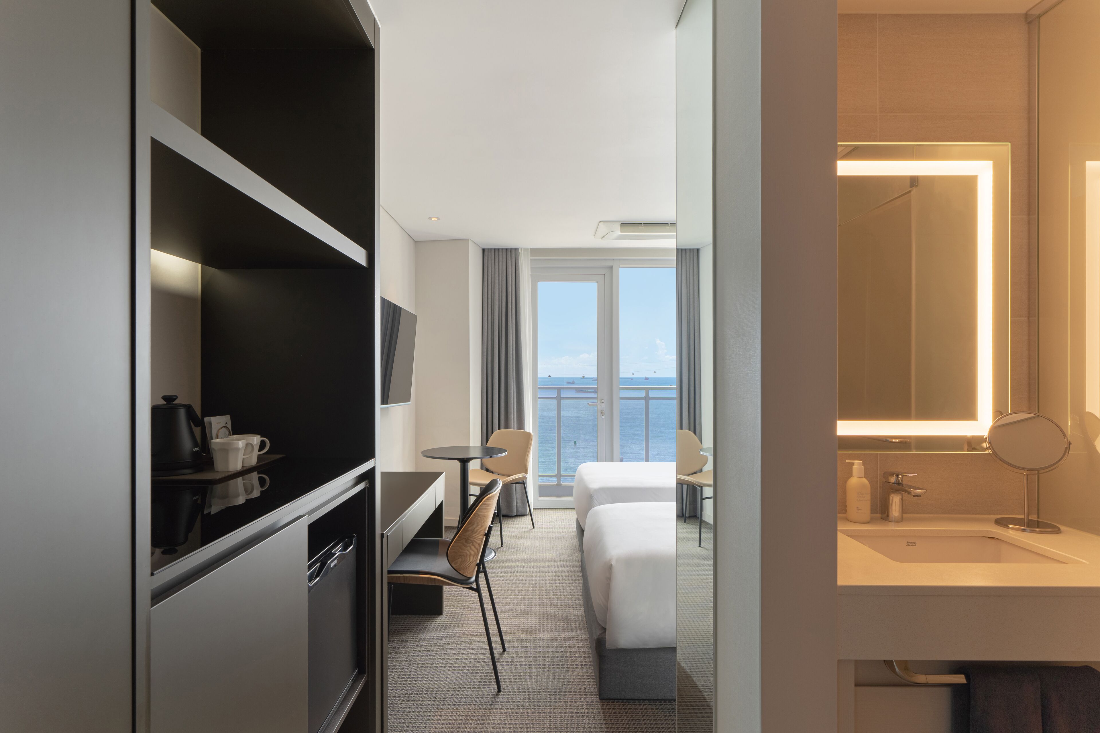 A hotel room with a bed, a desk, a chair, a TV, a bathroom with a mirror, and a view of the sea.