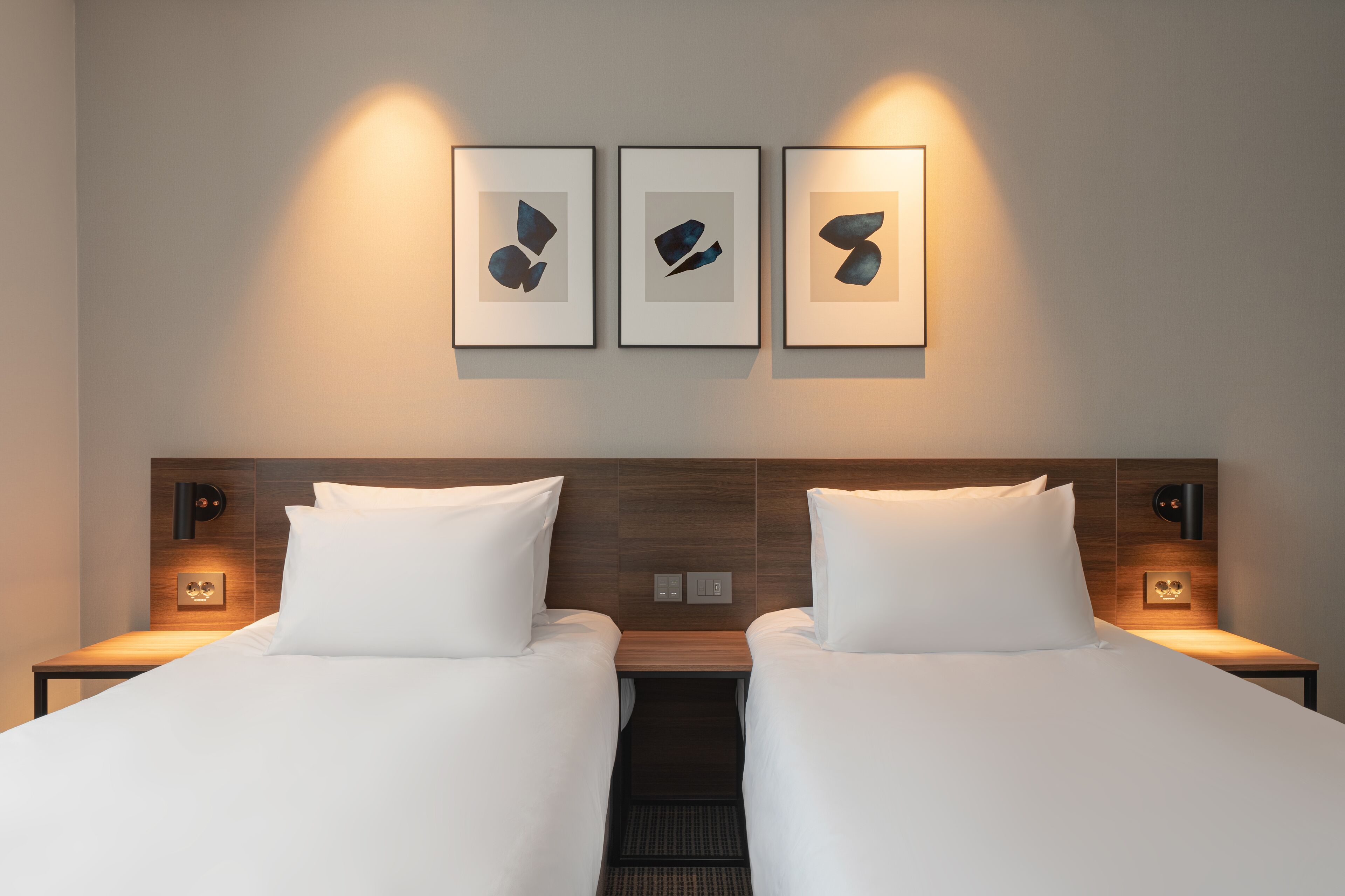 A hotel room with two beds, a wooden headboard, bedside tables with lamps, and three framed abstract artworks on the wall.