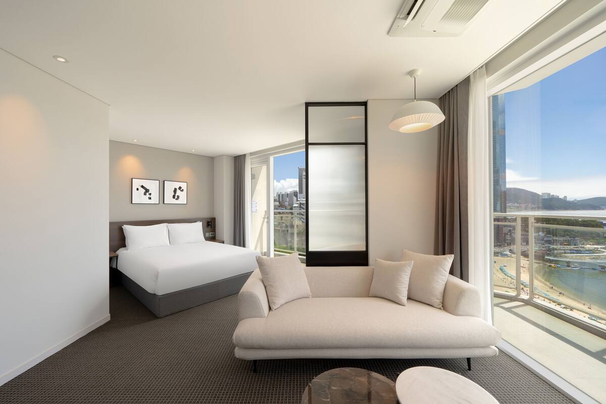 A modern hotel room with a bed, a sofa, a large window with a city view, and a mounted artwork on the wall.