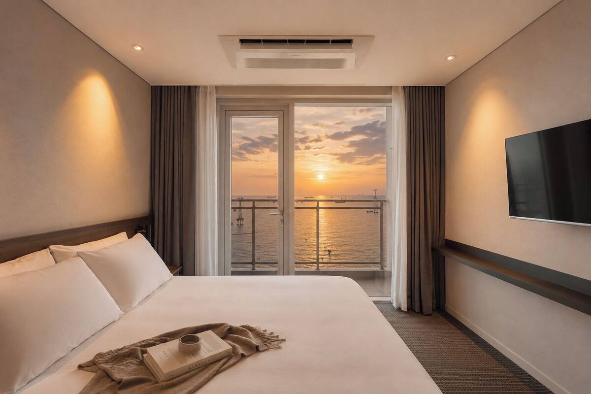 A hotel room with a bed, a flat-screen TV, a balcony with a sunset view, and an air conditioning unit.