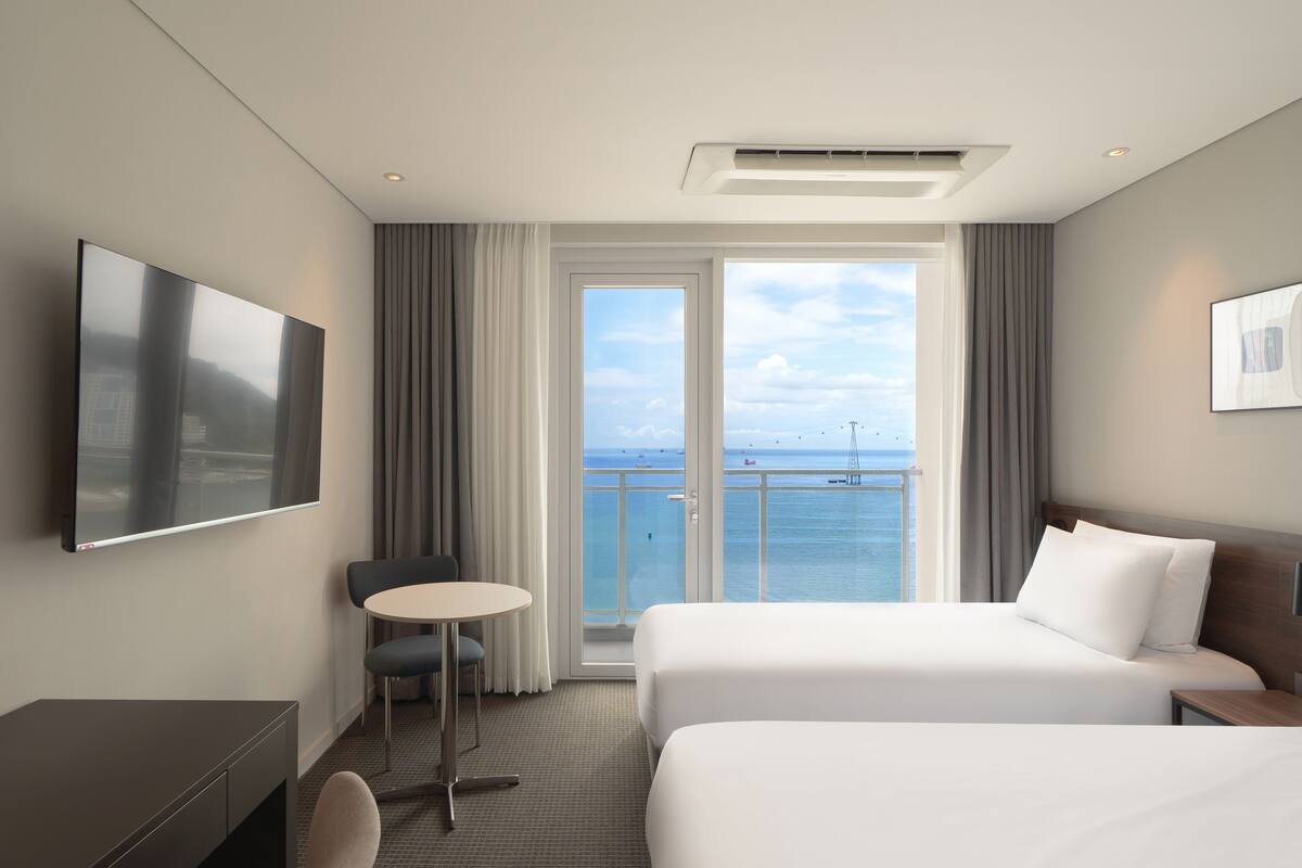A hotel room with a bed, a desk, a chair, a TV, and a balcony with a view of the sea.