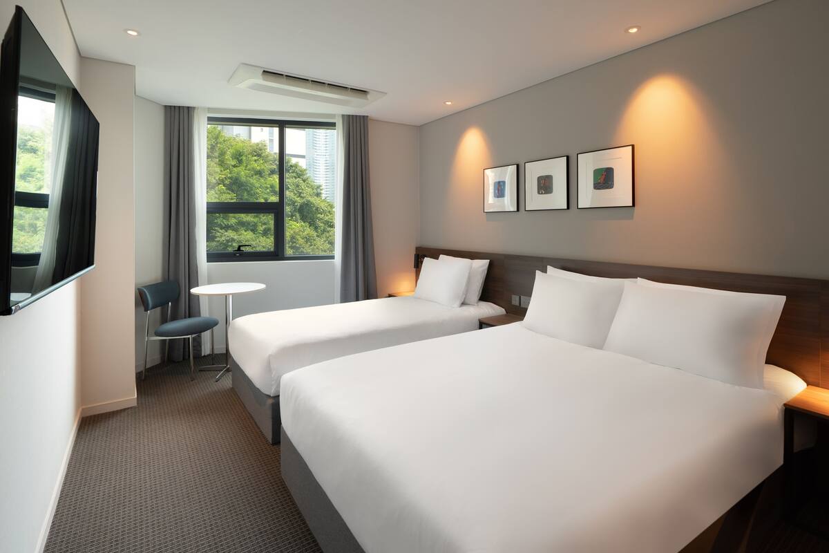 A hotel room with two beds, a TV, a small table, and a window with a view of trees.