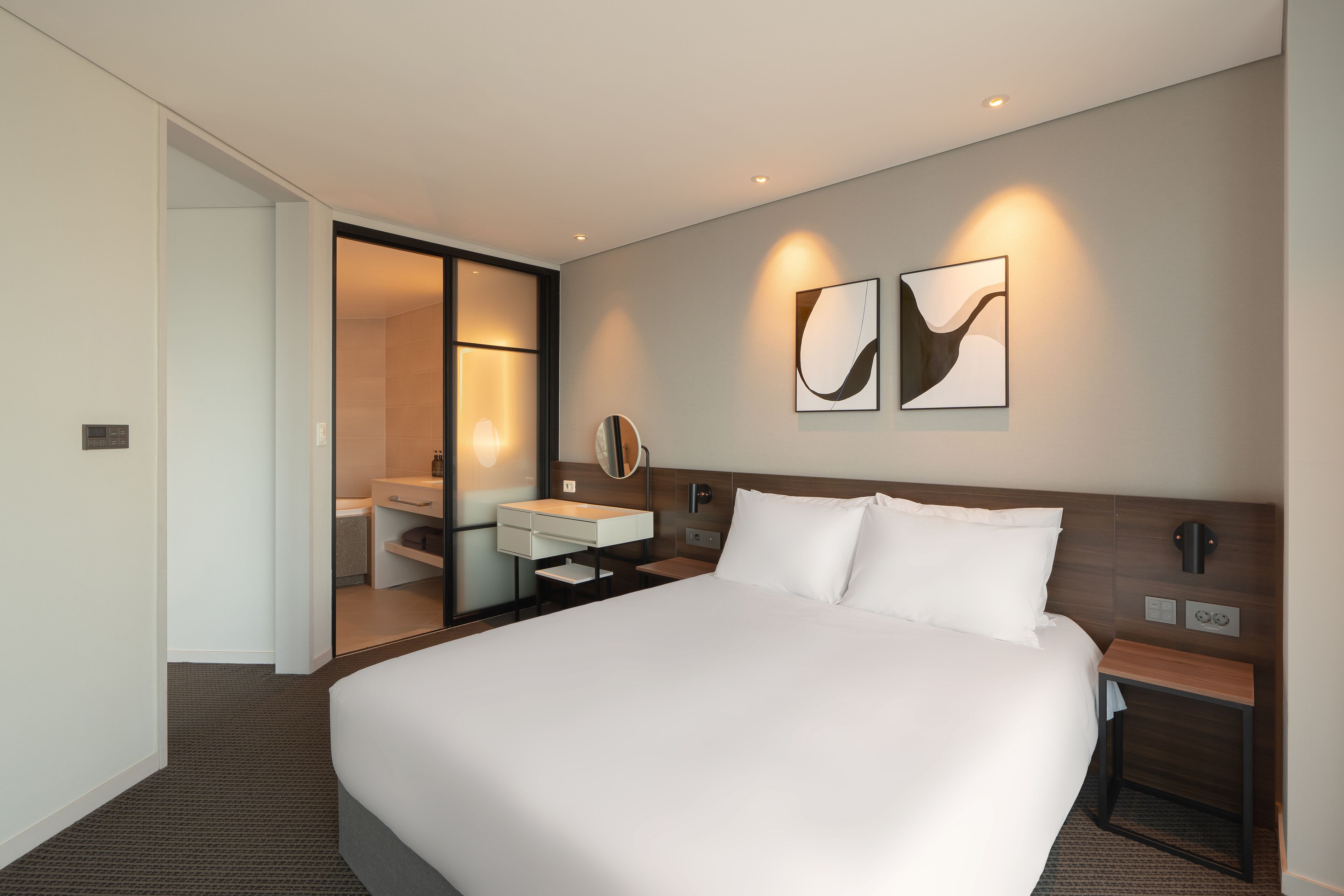 A modern hotel room with a large bed, a bedside table, a mirror, and a wall-mounted artwork.