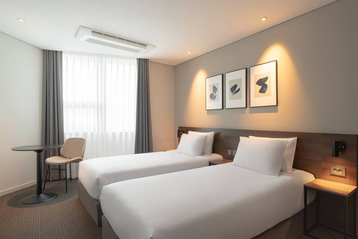 A hotel room with two beds, a small table, a chair, and three framed pictures on the wall.