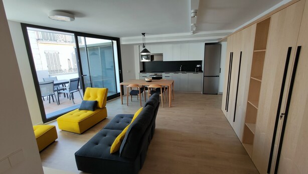Un apartamento moderno con sala de estar que incluye un sofá y una silla amarilla, una mesa de comedor con sillas, una cocina con armarios blancos y un ventanal corredizo que da a un balcón exterior.